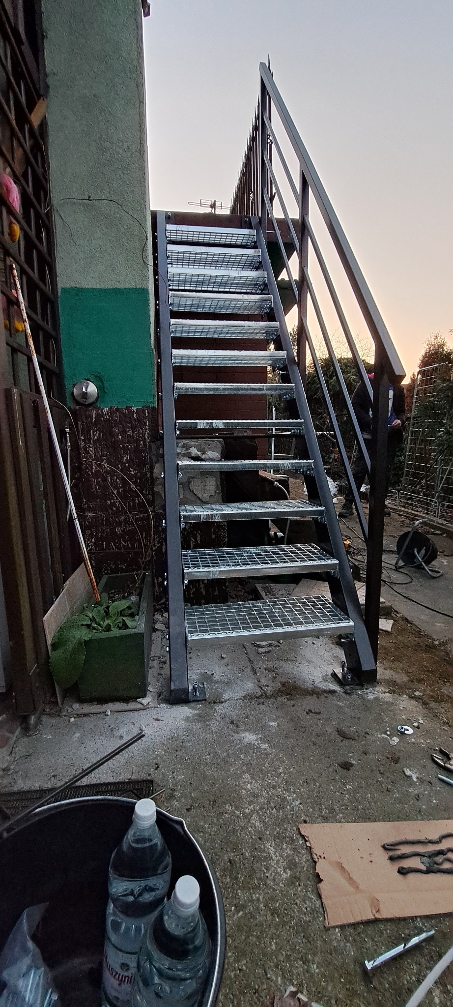 Zewnętrzne schody metalowe z kratowymi stopniami prowadzące na wyższy poziom, widoczne narzędzia i materiały montażowe na betonowym podłożu.