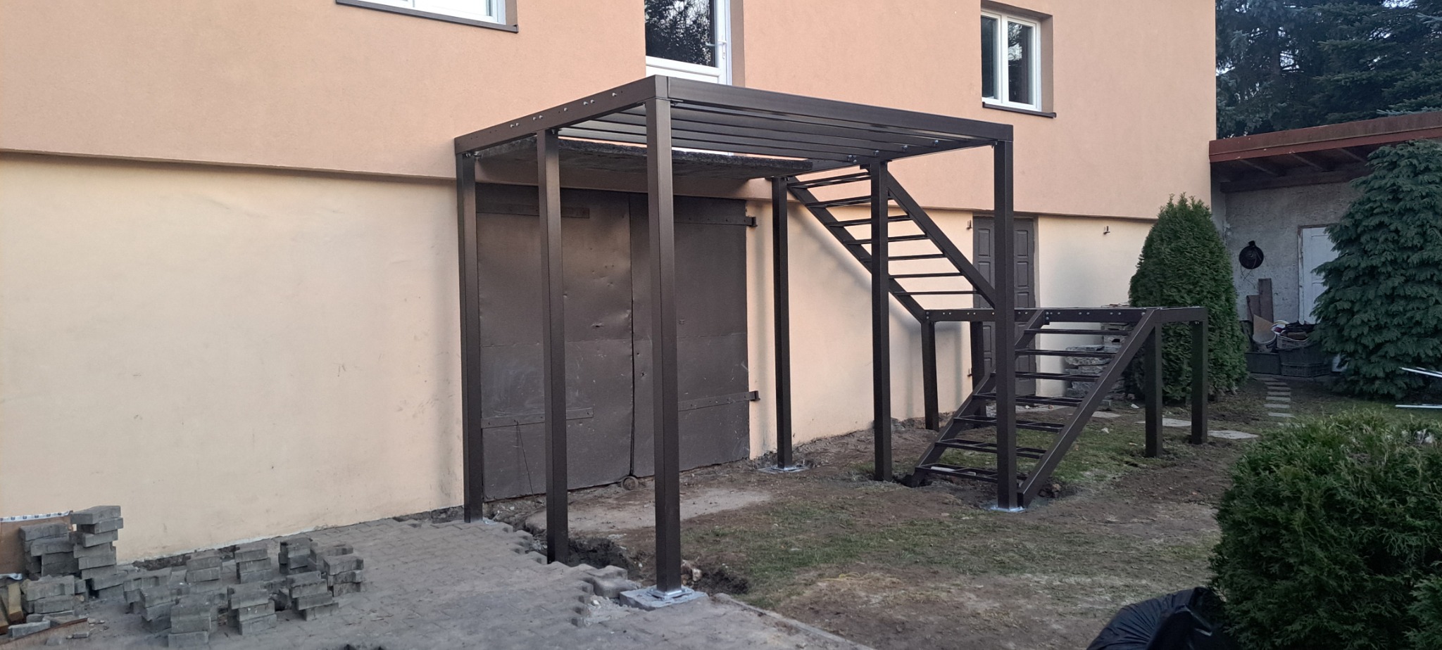 Zadaszone metalowe schody zewnętrzne prowadzące do piwnicy, widoczne kostki brukowe przygotowane do ułożenia wokół konstrukcji.