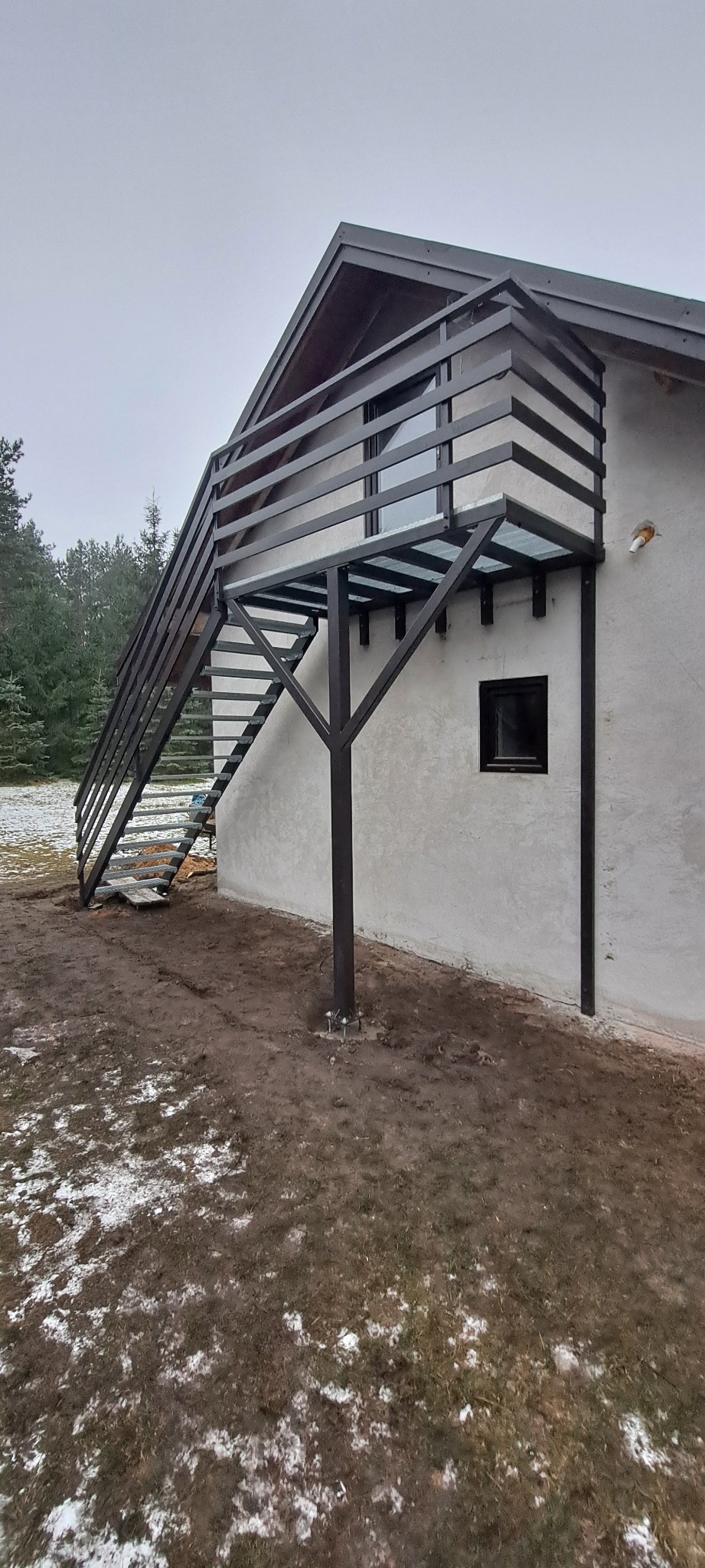 Metalowe schody zewnętrzne z balustradą prowadzące na balkon domu, widoczne na tle pochmurnego nieba i częściowo ośnieżonego terenu.