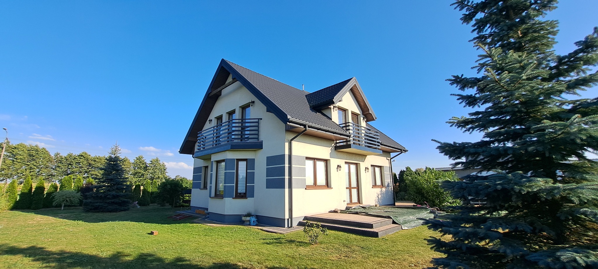 Dwukondygnacyjny dom z balkonami z prostymi, metalowymi barierkami, widoczny z perspektywy ogrodu w słoneczny dzień. Elewacja w odcieniach beżu i szarości.