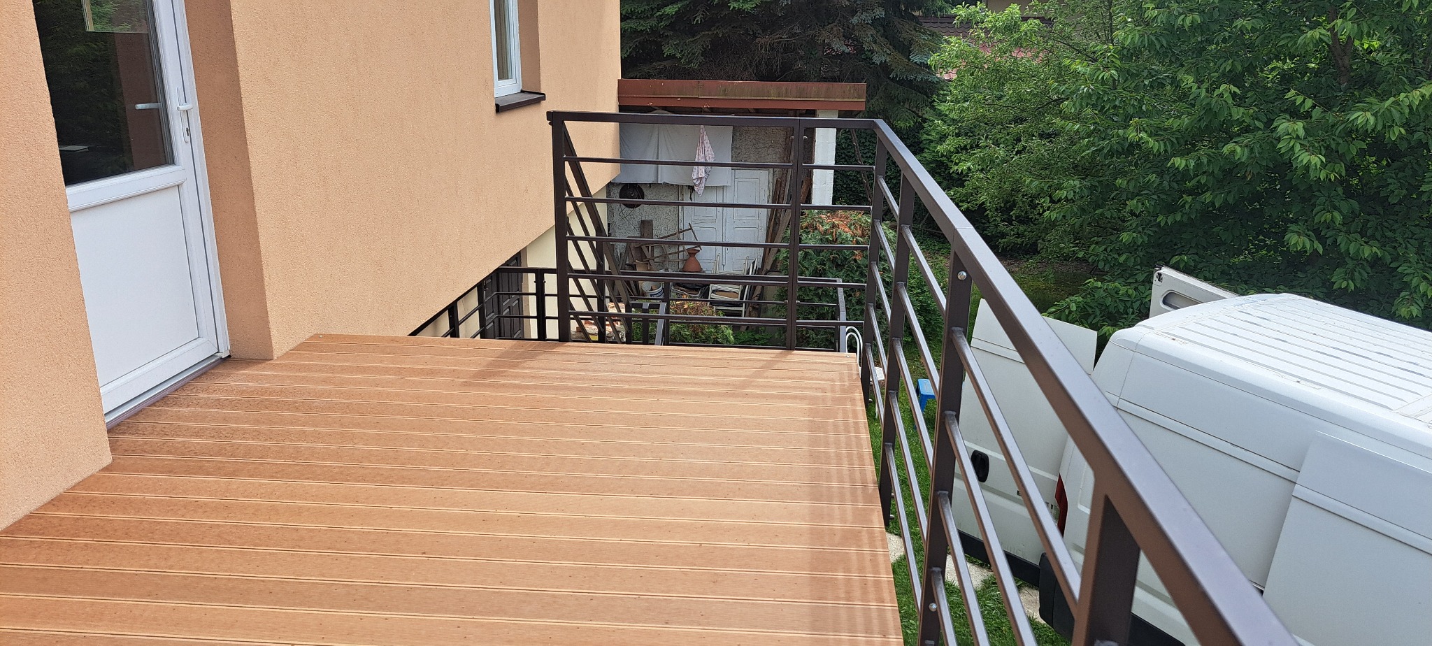 Balkon z drewnianą podłogą i metalową, ciemną balustradą. Widok z góry na fragment balkonu, zieleń w tle i biały samochód dostawczy.