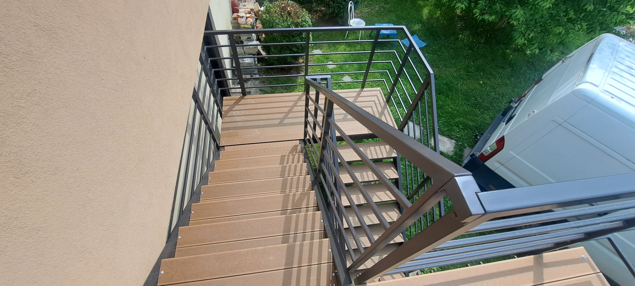 Zewnętrzne schody z brązowymi deskami kompozytowymi i ciemną, metalową balustradą, widziane z góry. Obok zaparkowany biały bus.