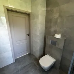 Nowoczesna łazienka z szarą płytką, podwieszaną toaletą i rolką papieru toaletowego na zabudowie stelaża; drzwi w odcieniu złamanej bieli z prostą klamką.