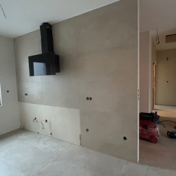 NS Wykończenia Wnętrz - Wnętrze w trakcie prac wykończeniowych: zamontowany czarny okap kuchenny, częściowo położone płytki na ścianie, widoczne otwory pod instalacje elektryczne, podłoga wyłożona płytkami, w tle...