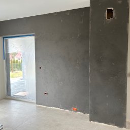 NS Wykończenia Wnętrz - Ściana w surowym, szarym tynku dekoracyjnym z widocznymi śladami szpachlowania, z przygotowanymi otworami na gniazdka elektryczne i puszką instalacyjną, zabezpieczone okno folią malarską, jasna...