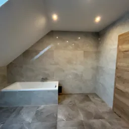 Wykończona łazienka na poddaszu z wanną obudowaną płytkami w odcieniach szarości, ściany wyłożone płytkami imitującymi marmur, drewniane drzwi i punktowe oświetlenie sufitowe.
