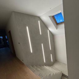 NS Wykończenia Wnętrz - Wnętrze w trakcie remontu: szare ściany z pionowymi paskami ledowego oświetlenia, betonowe schody prowadzące w dół, drewniana podłoga na pierwszym planie, okno dachowe z widokiem na niebo.