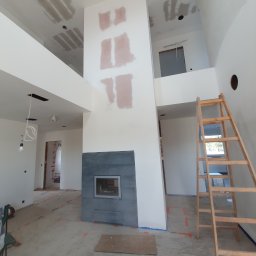 NS Wykończenia Wnętrz - Wnętrze domu w trakcie prac wykończeniowych: surowy kominek z ciemnej płytki, ściany z widocznymi śladami szpachlowania, drewniana drabina oparta o ścianę, zabezpieczona podłoga.