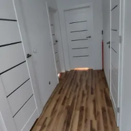 Korytarz z nową podłogą z paneli imitujących drewno, białe ściany i trzy białe drzwi z poziomymi czarnymi wstawkami, widoczny właz na suficie.