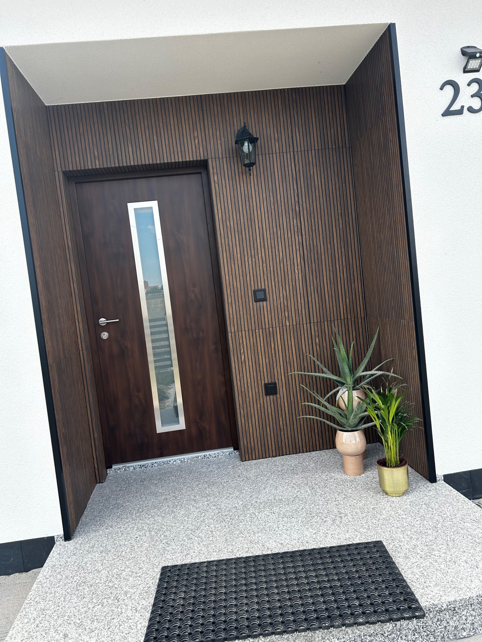 Nowoczesne wejście do domu z brązowymi panelami ściennymi, drzwiami z pionowym przeszkleniem, numerem domu '23', oświetleniem zewnętrznym i roślinami w donicach na szarym podłożu z czarną wycieraczką.
