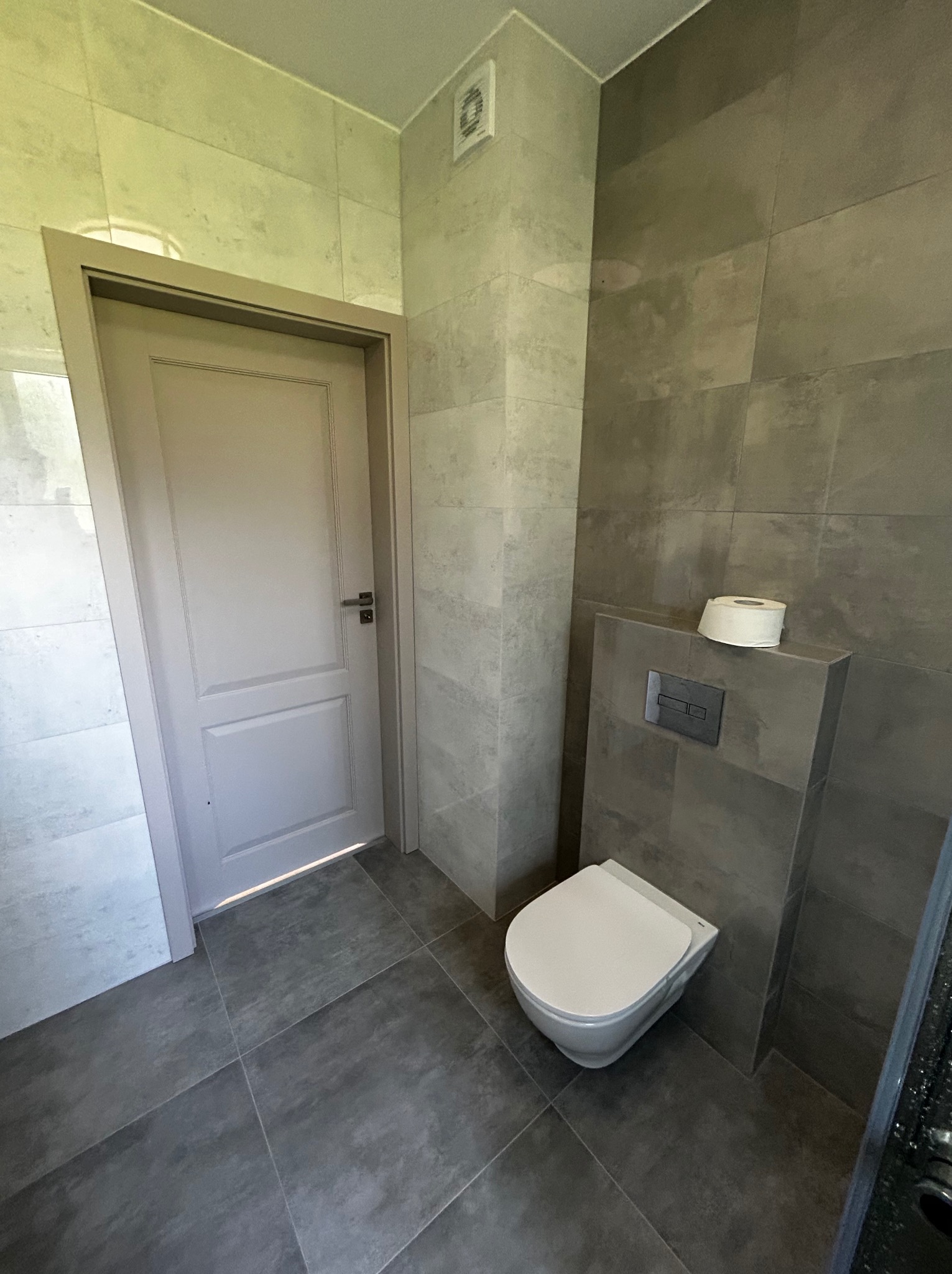 Nowoczesna łazienka z szarą płytką, podwieszaną toaletą i rolką papieru toaletowego na zabudowie stelaża; drzwi w odcieniu złamanej bieli z prostą klamką.