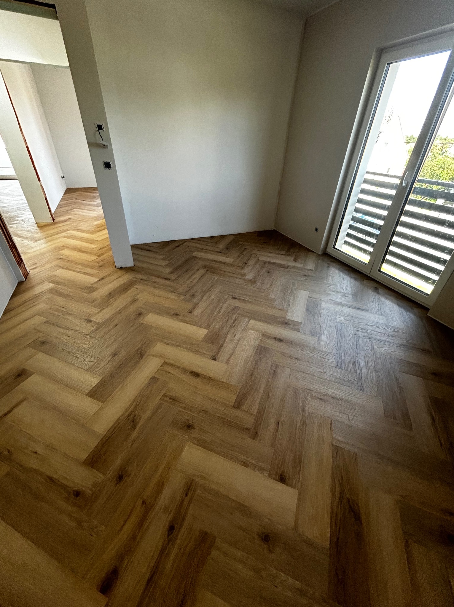 Jasne panele podłogowe ułożone w jodełkę francuską w nowym pokoju z białymi ścianami i oknem balkonowym, widok na zewnątrz.