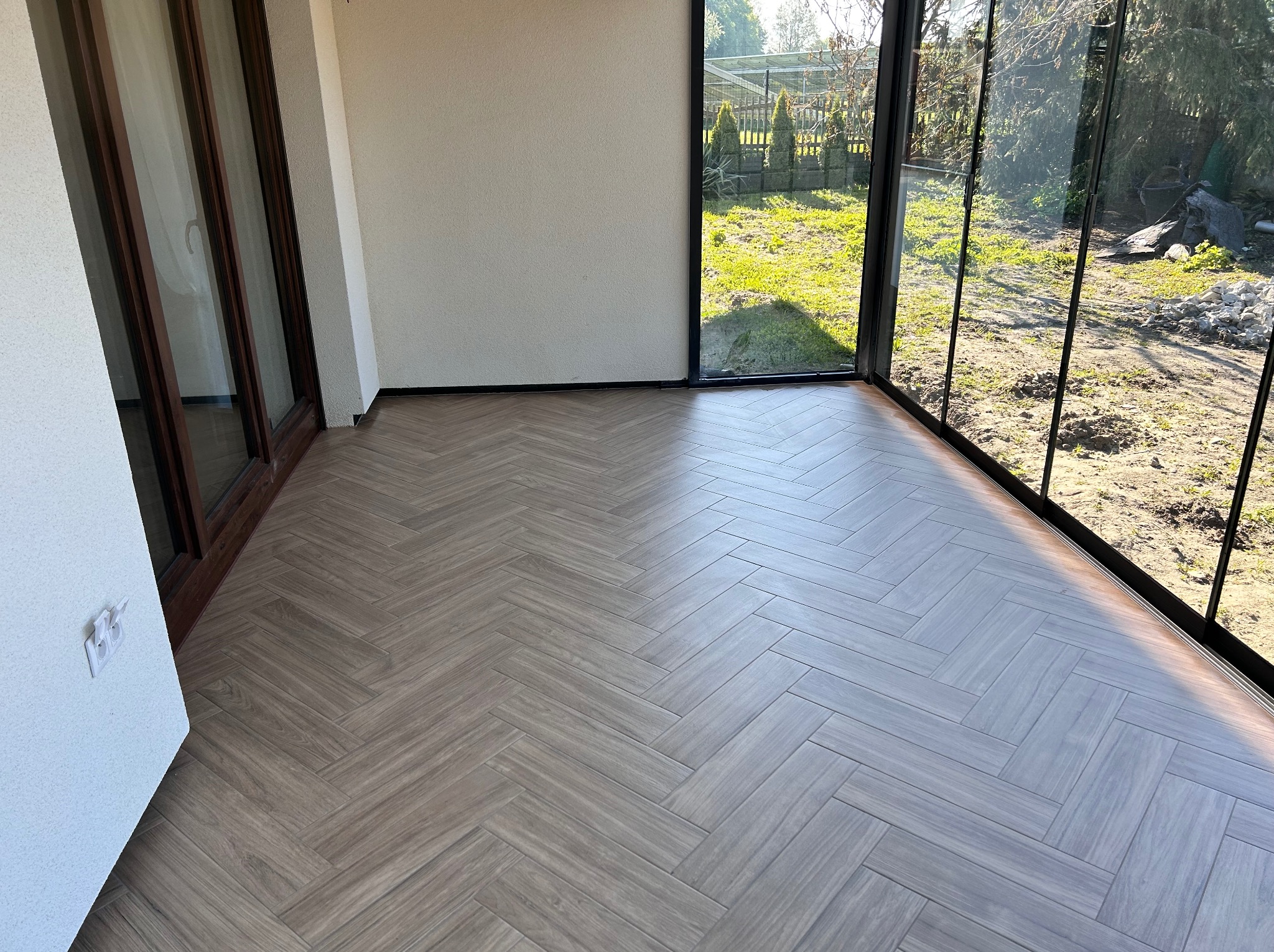 Ułożona jodełka z paneli podłogowych w jasnym odcieniu drewna, widok na ogród przez przeszkloną ścianę, gniazdko elektryczne na białej ścianie po lewej.