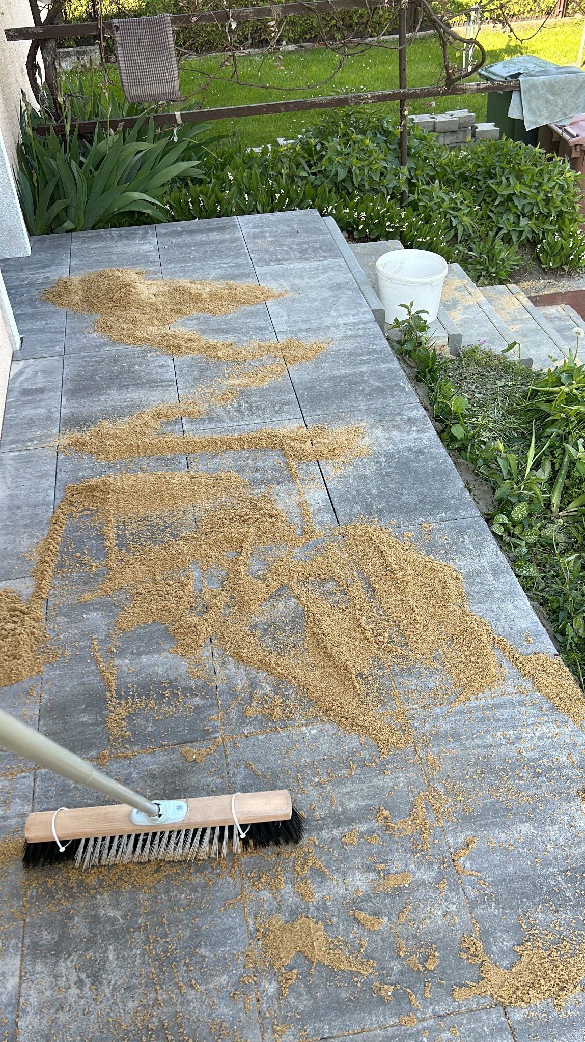 Szczotka zamiata piasek na szarych płytach chodnikowych przed domem, widoczny fragment ogrodu w tle.