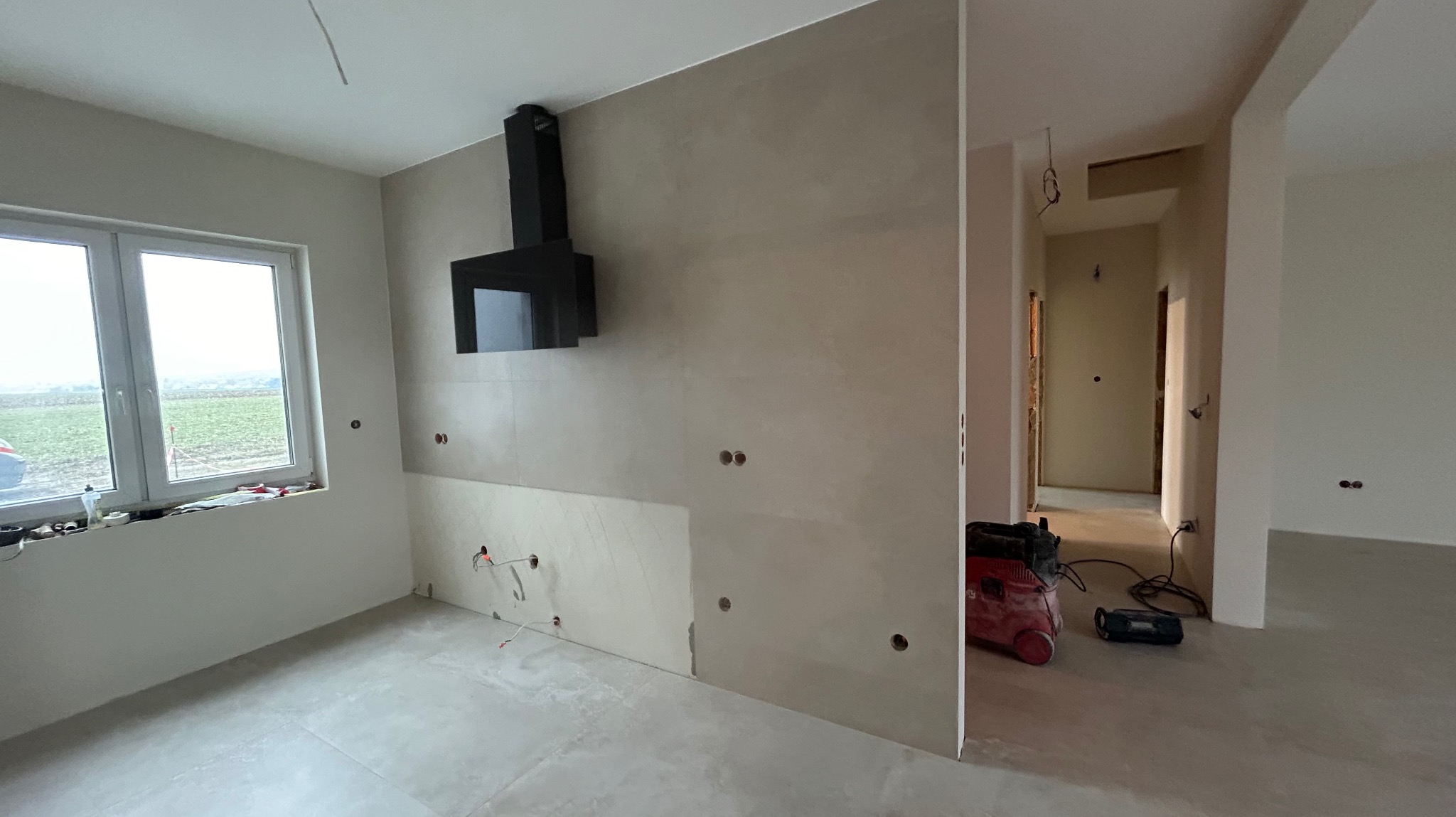 Wnętrze w trakcie prac wykończeniowych: zamontowany czarny okap kuchenny, częściowo położone płytki na ścianie, widoczne otwory pod instalacje elektryczne, podłoga wyłożona płytkami, w tle...