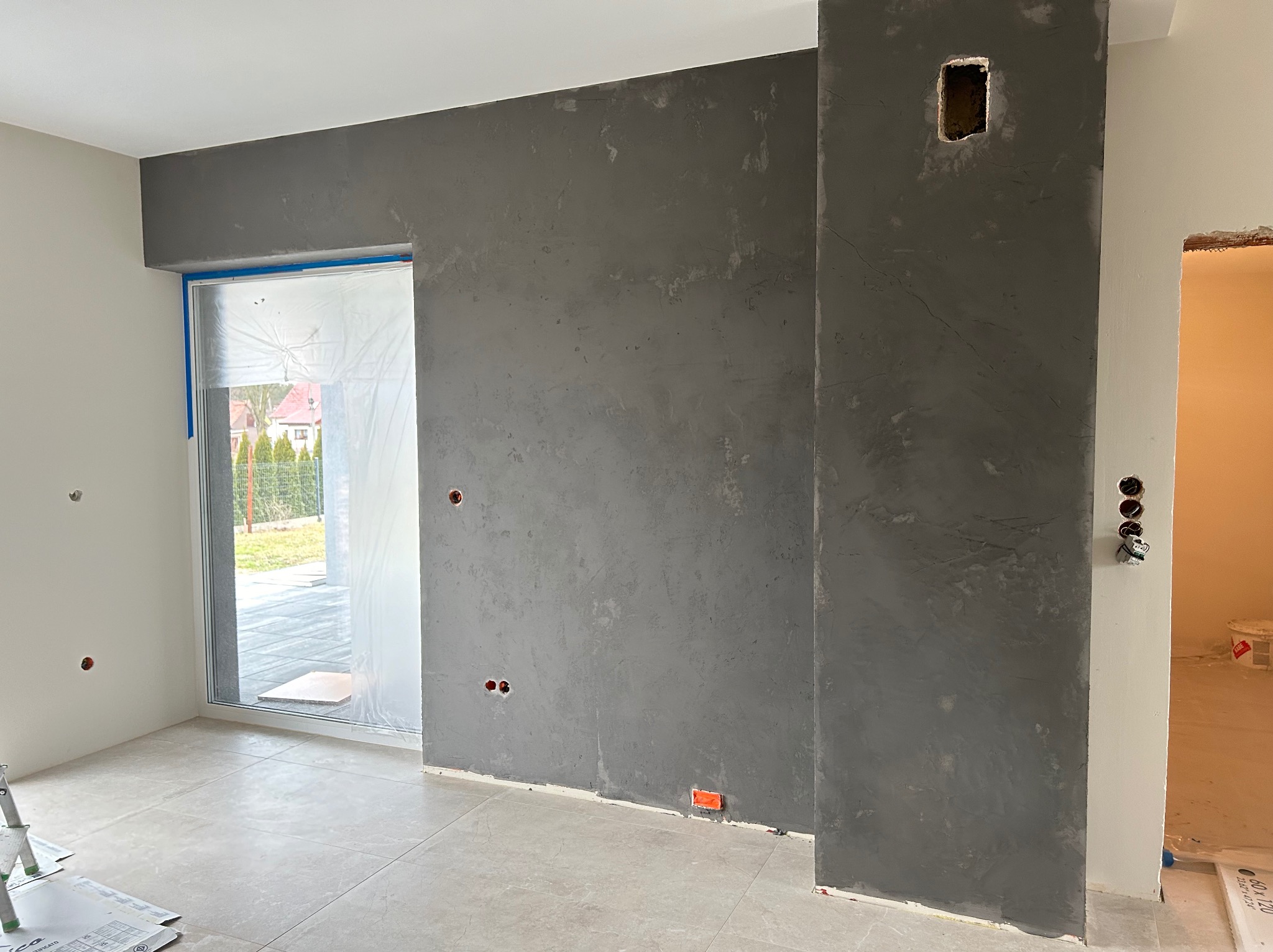 Ściana w surowym, szarym tynku dekoracyjnym z widocznymi śladami szpachlowania, z przygotowanymi otworami na gniazdka elektryczne i puszką instalacyjną, zabezpieczone okno folią malarską, jasna...