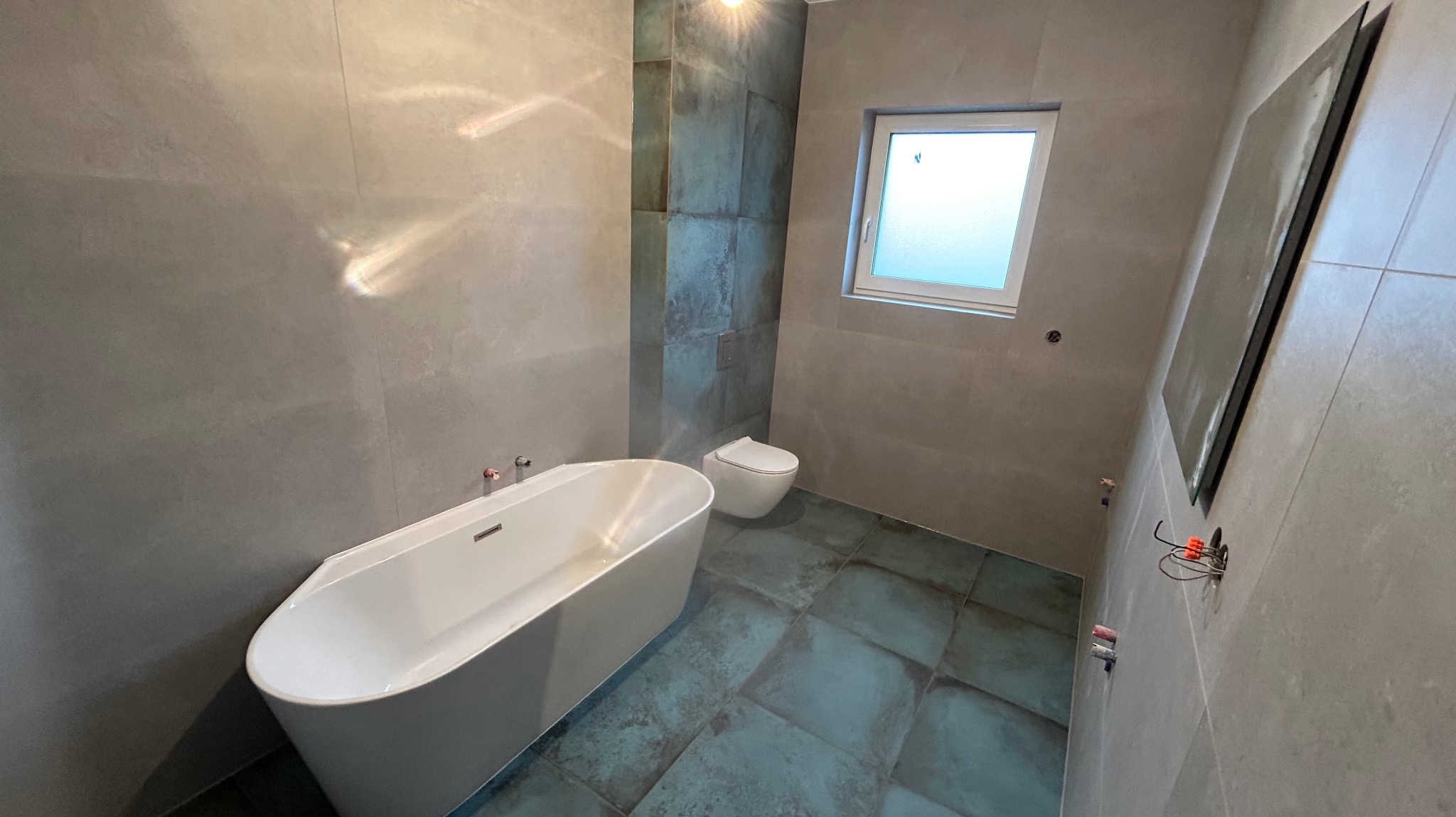 Nowoczesna łazienka z wolnostojącą białą wanną, podwieszaną toaletą i dużym lustrem na szarej ścianie z płytek, akcentowana kolumną z turkusowych płytek.