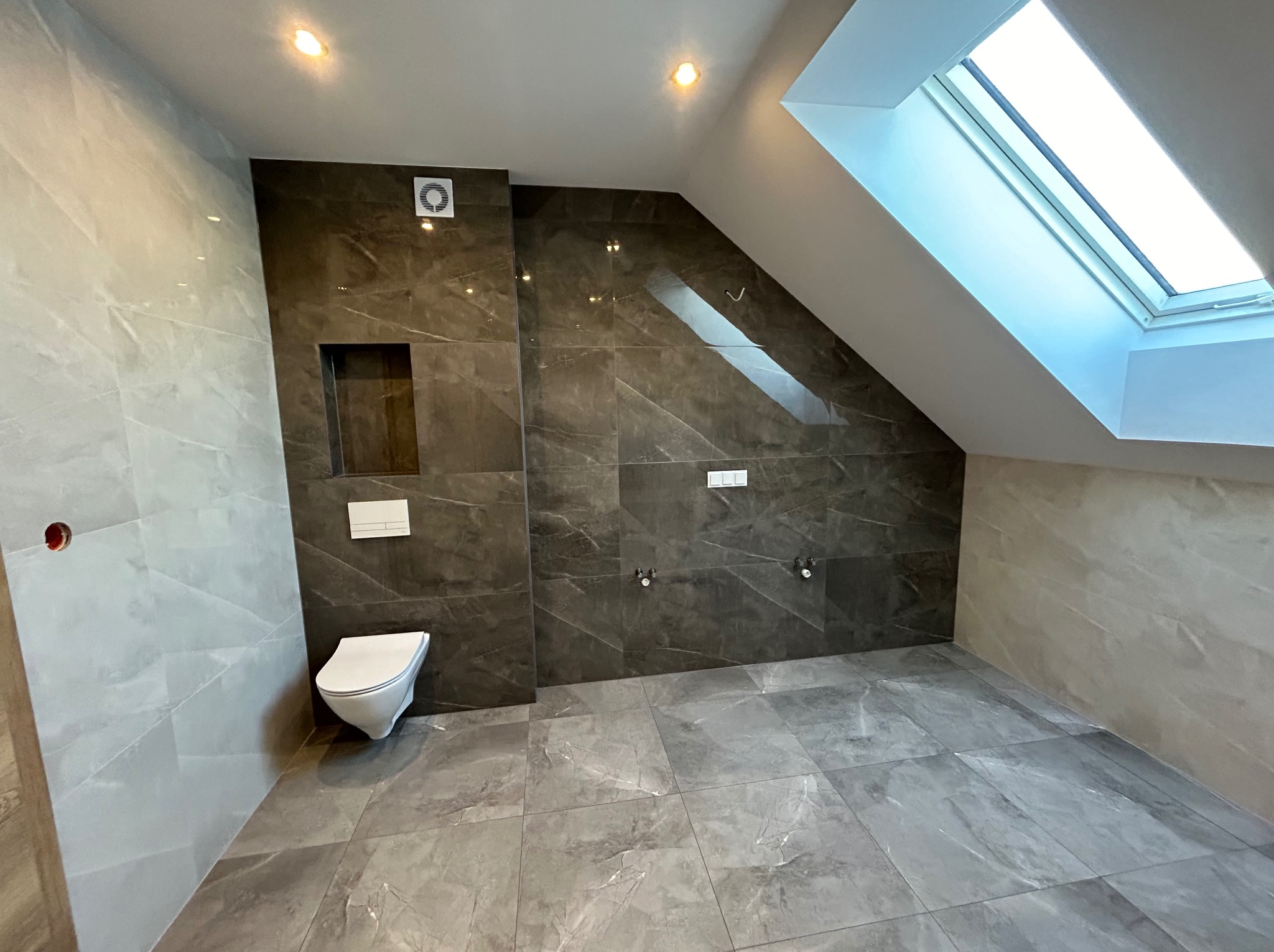 Wykończona łazienka na poddaszu z szarymi płytkami imitującymi marmur na ścianach i podłodze, wisząca toaleta, okno dachowe wpuszczające dużo światła.
