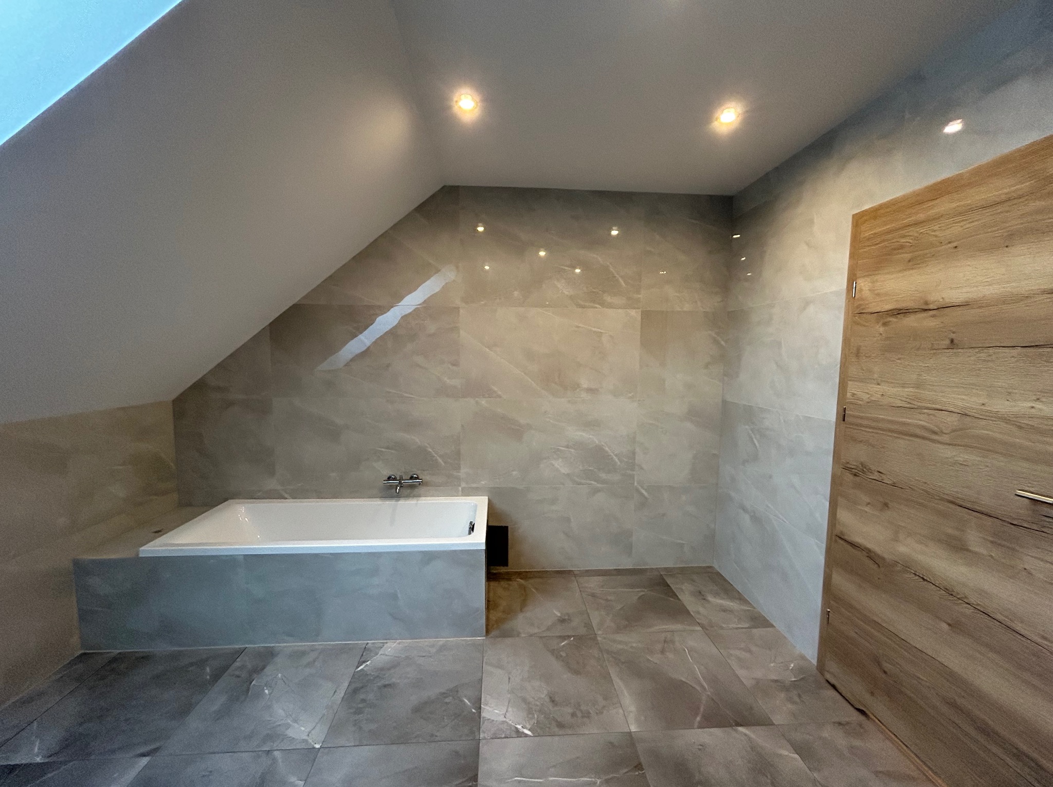 Wykończona łazienka na poddaszu z wanną obudowaną płytkami w odcieniach szarości, ściany wyłożone płytkami imitującymi marmur, drewniane drzwi i punktowe oświetlenie sufitowe.