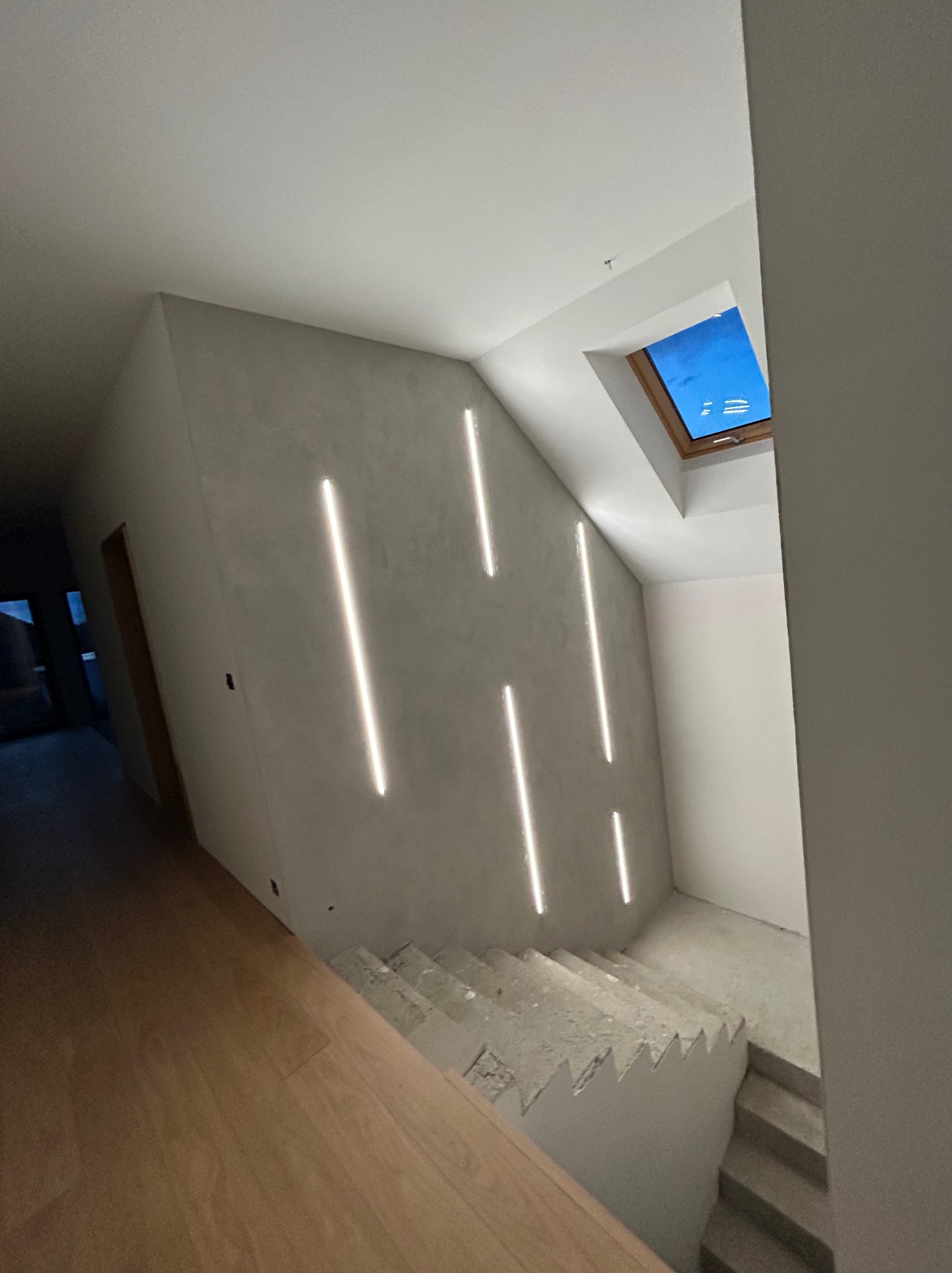 Wnętrze w trakcie remontu: szare ściany z pionowymi paskami ledowego oświetlenia, betonowe schody prowadzące w dół, drewniana podłoga na pierwszym planie, okno dachowe z widokiem na niebo.