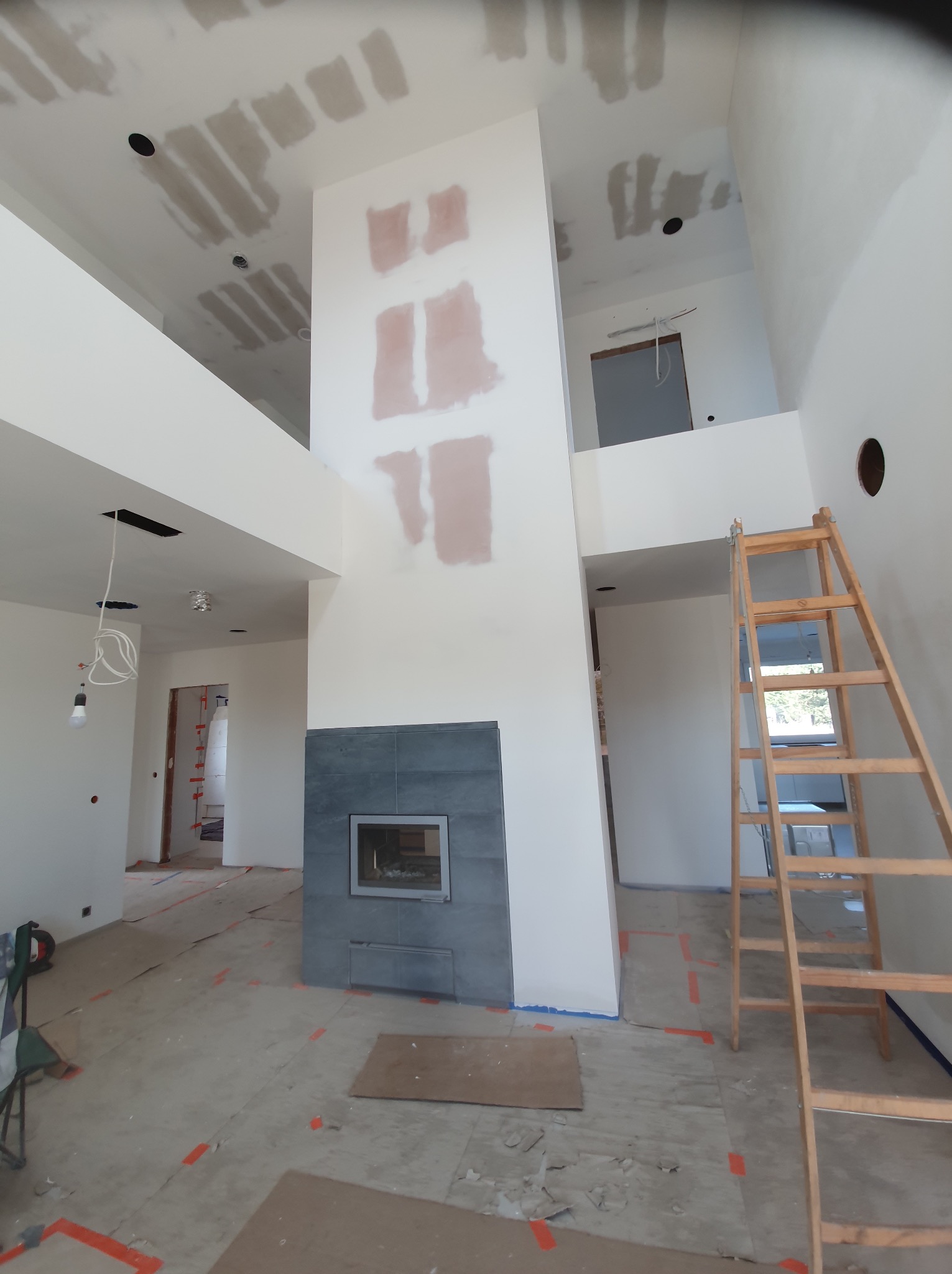 Wnętrze domu w trakcie prac wykończeniowych: surowy kominek z ciemnej płytki, ściany z widocznymi śladami szpachlowania, drewniana drabina oparta o ścianę, zabezpieczona podłoga.