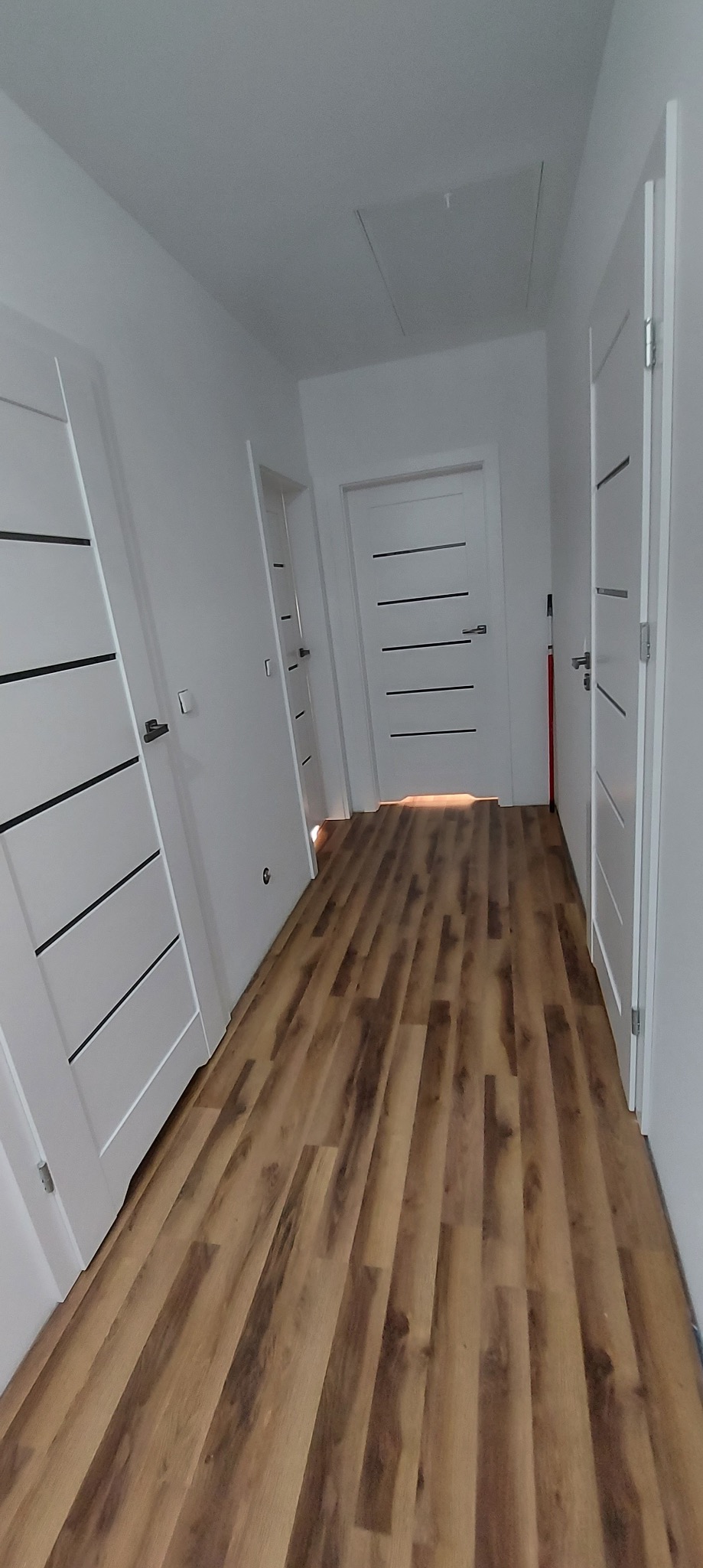 Korytarz z nową podłogą z paneli imitujących drewno, białe ściany i trzy białe drzwi z poziomymi czarnymi wstawkami, widoczny właz na suficie.