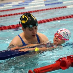 Oliwia Król - Instruktor w czapce z logo Batmana uczy dziewczynkę z różowymi okularami i deską pływacką w basenie z czerwonymi torami.