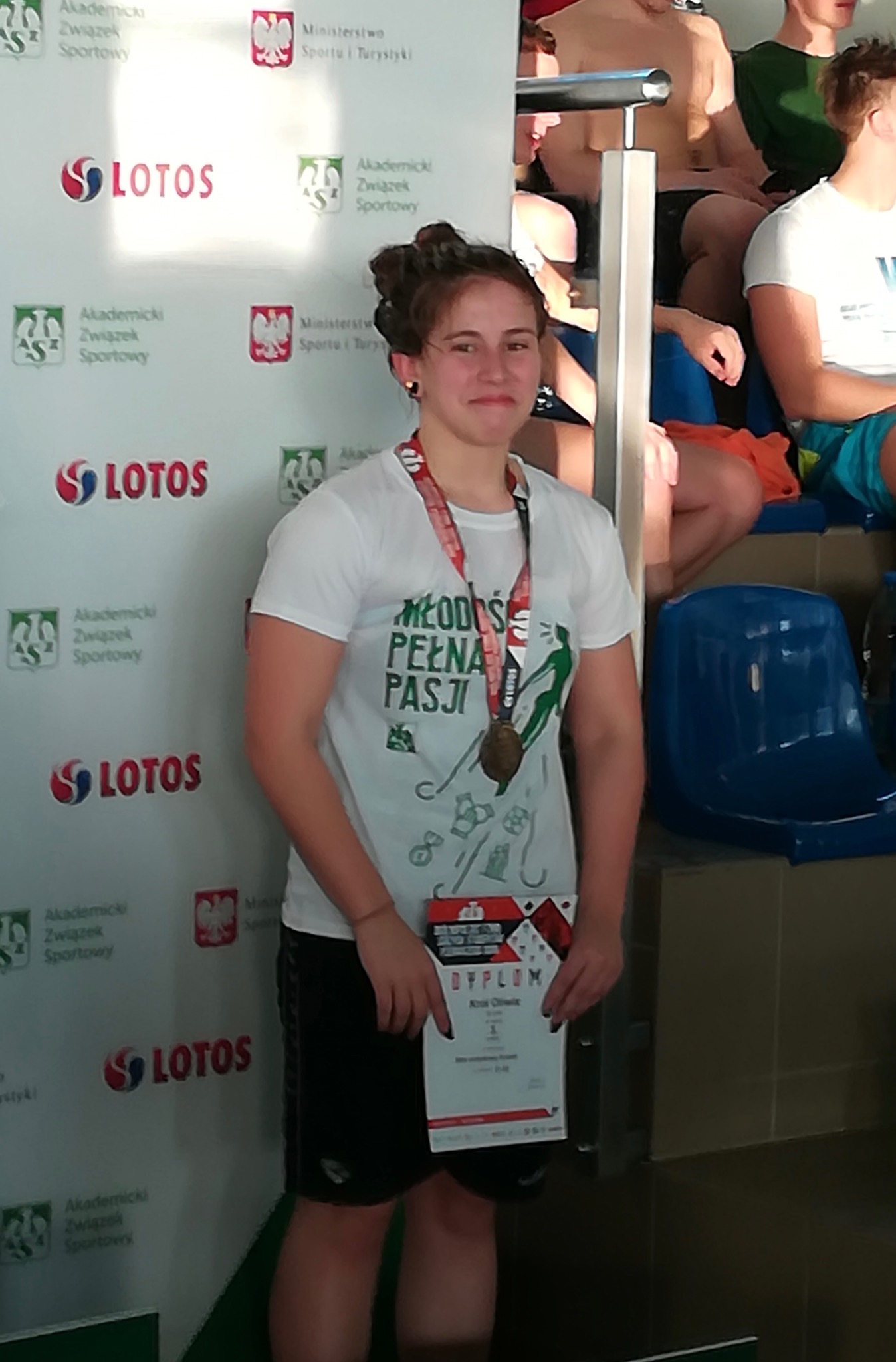 Młoda kobieta z medalem na szyi i dyplomem w ręku, na tle ściany z logo LOTOS i Akademii Związku Sportowego.