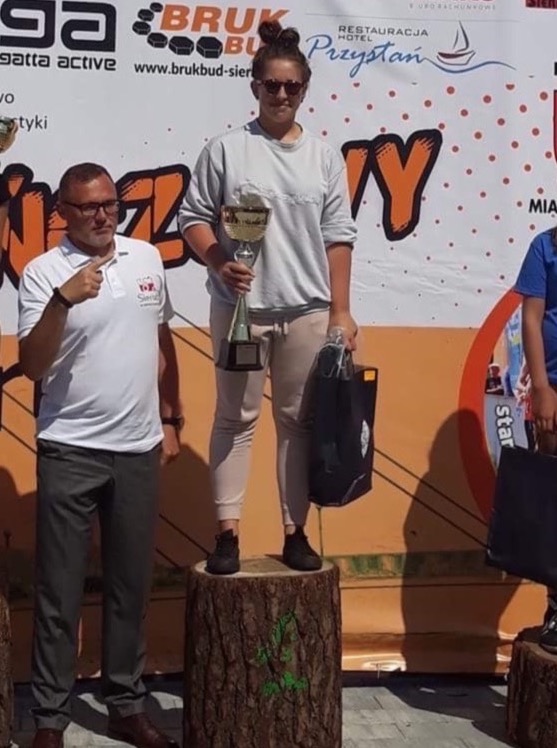 Zwyciężczyni zawodów sportowych stoi na drewnianym podium, trzymając puchar i torbę, obok niej mężczyzna w koszulce polo oraz fragment sylwetki dziecka.