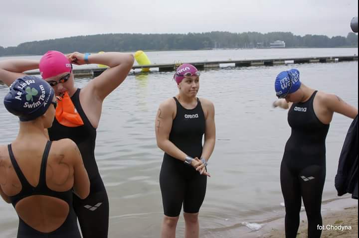 Grupa zawodniczek w strojach pływackich Arena i czepkach, stojąca przy brzegu jeziora przed zawodami, widoczne boje na wodzie i pomost w tle.