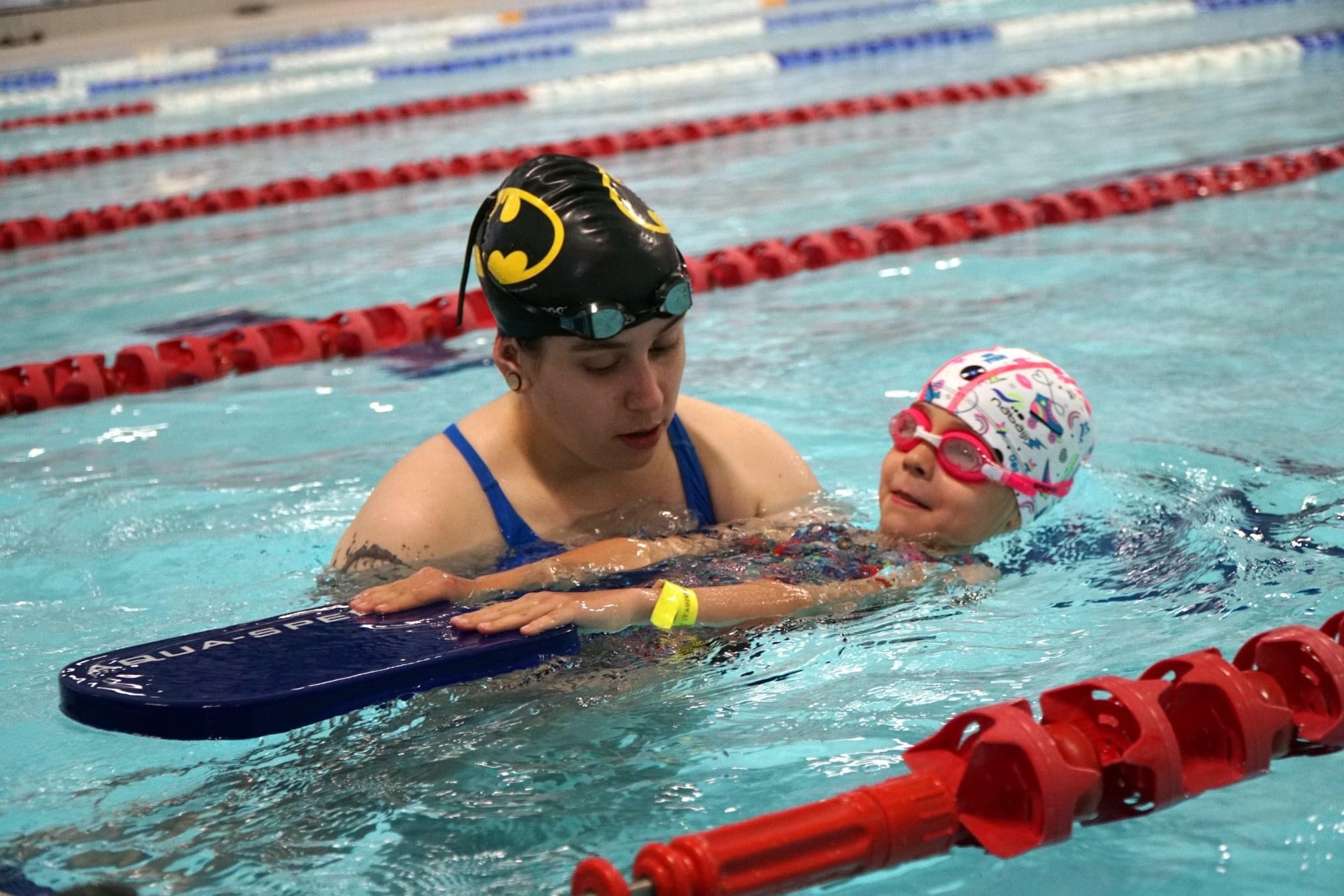 Instruktor w czapce z logo Batmana uczy dziewczynkę z różowymi okularami i deską pływacką w basenie z czerwonymi torami.