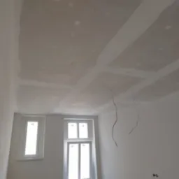 Sufit z płyt karton gips, szpachlowany, z widocznymi przewodami elektrycznymi i dwoma oknami w białych ramach.