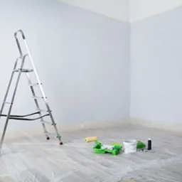 Pokój w trakcie malowania na szaro, zabezpieczona podłoga folią, drabina aluminiowa, wałek i kuweta malarska, pędzel. Widok na róg pokoju.