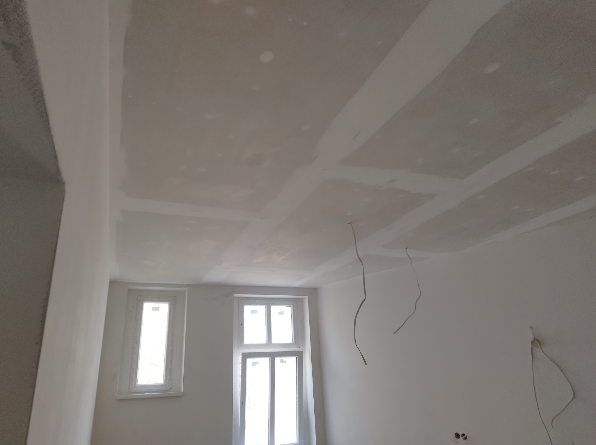 Sufit z płyt karton gips, szpachlowany, z widocznymi przewodami elektrycznymi i dwoma oknami w białych ramach.