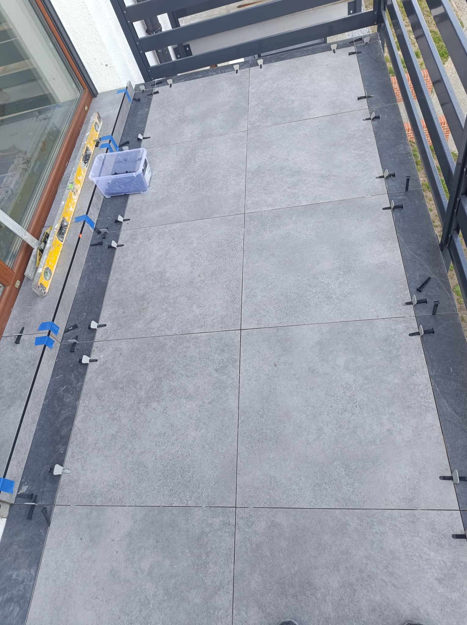 Montaż szarych płytek na tarasie z użyciem poziomicy, klipsów i klinów do wyrównywania, widoczna balustrada.