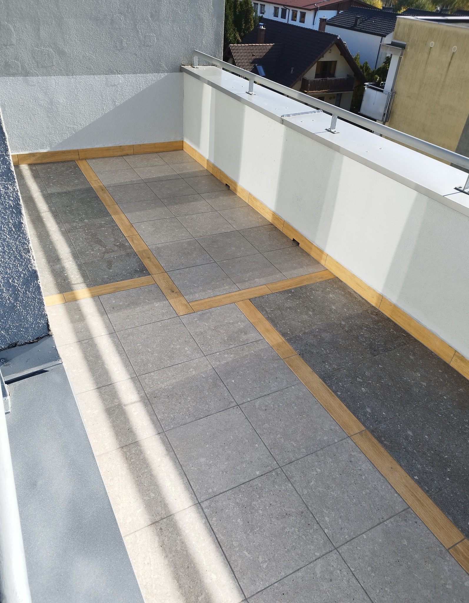 Wykończony taras na balkonie z szarych płytek z drewnianymi wstawkami, widok z góry na jasnym słońcu.