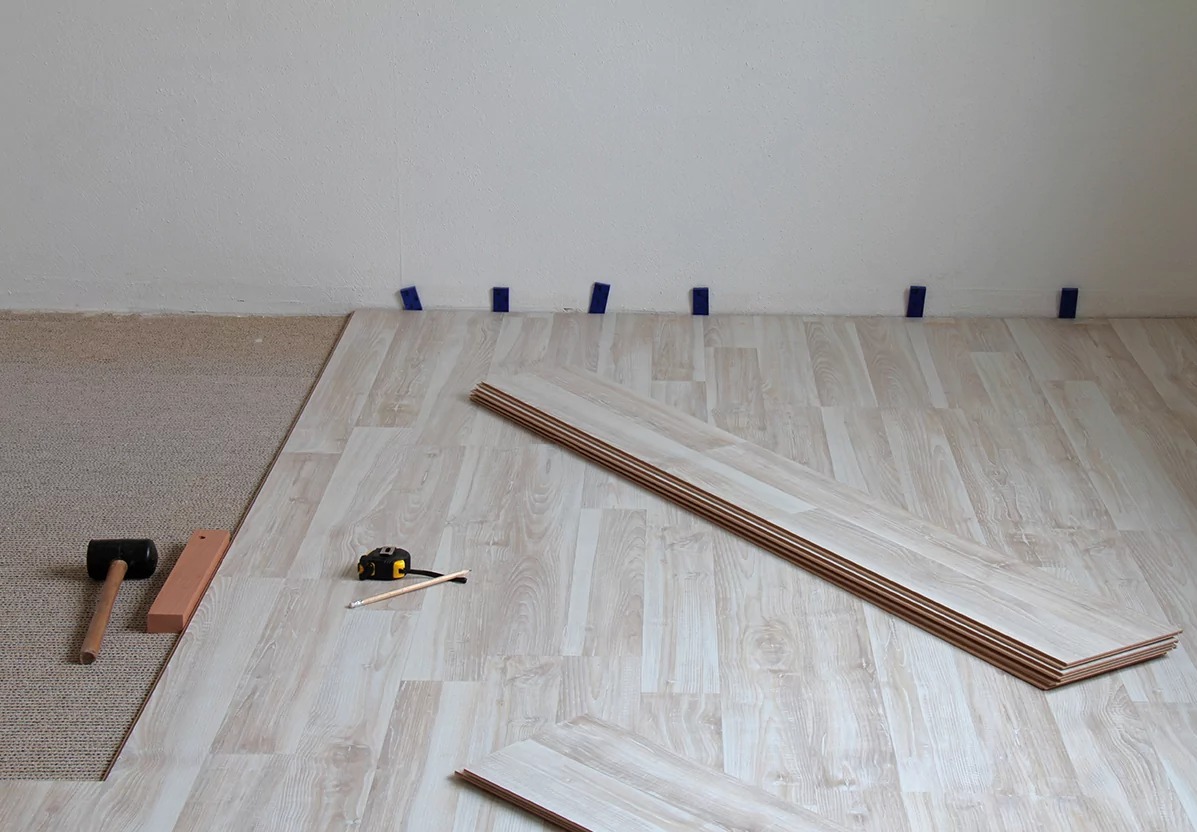 Fragment podłogi w trakcie układania paneli. Część podłogi pokryta wykładziną, na pozostałej części ułożone panele. Widoczne narzędzia: młotek, miarka, ołówek.