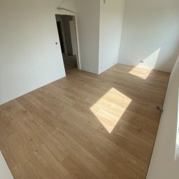 Jasne panele podłogowe w nowym pokoju z białymi ścianami, widok na otwarte drzwi do kolejnego pomieszczenia. Promienie słońca na podłodze.