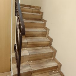 Zakład Remontowo Budowlany MACBUD - Schody wyłożone płytkami imitującymi drewno, z metalową balustradą, widoczne ślady po kleju i fugowaniu.