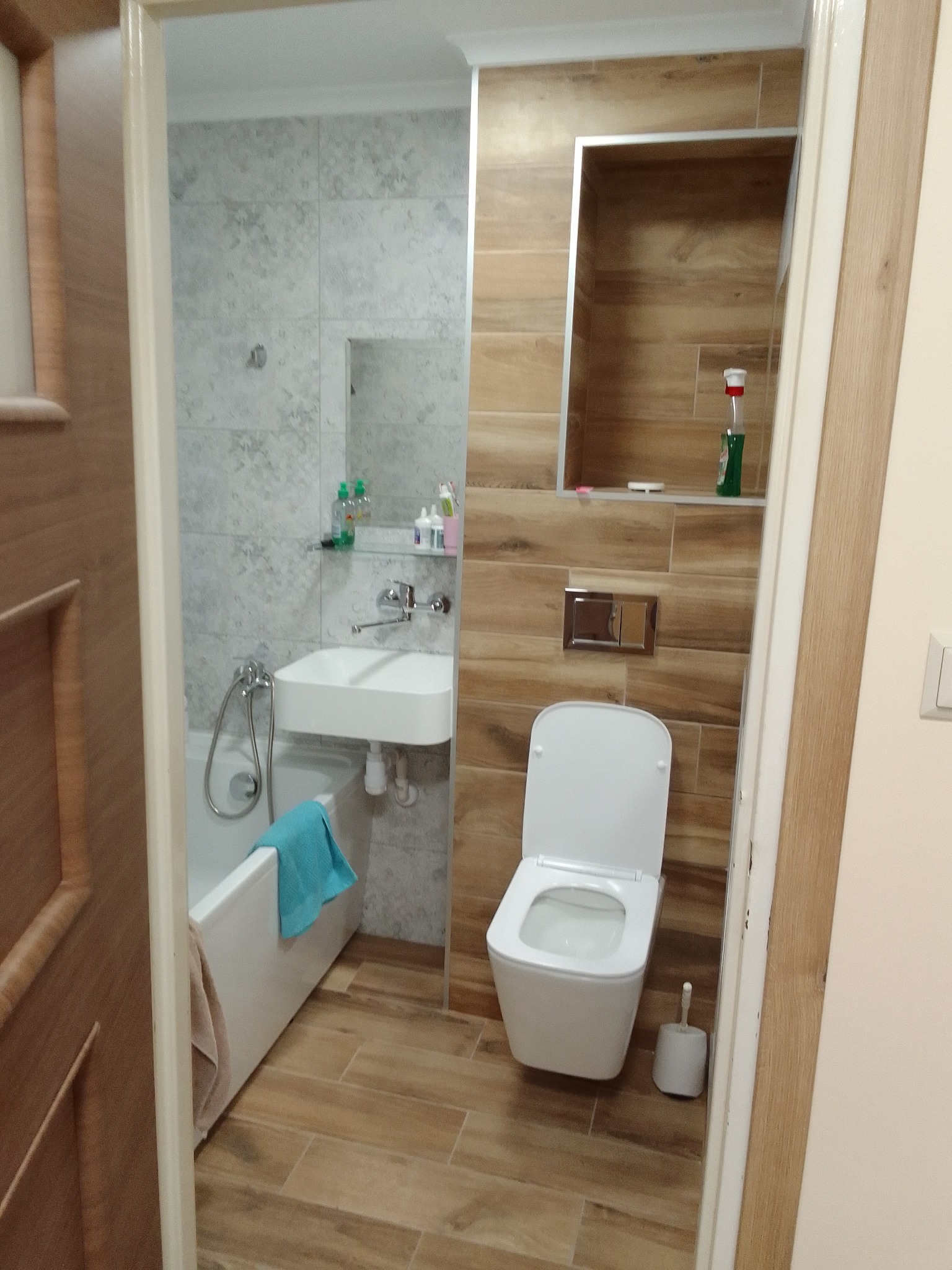 Wykończona łazienka z wanną, umywalką nablatową, toaletą podwieszaną, płytkami imitującymi drewno i szare płytki z wzorem, widziana z perspektywy drzwi.