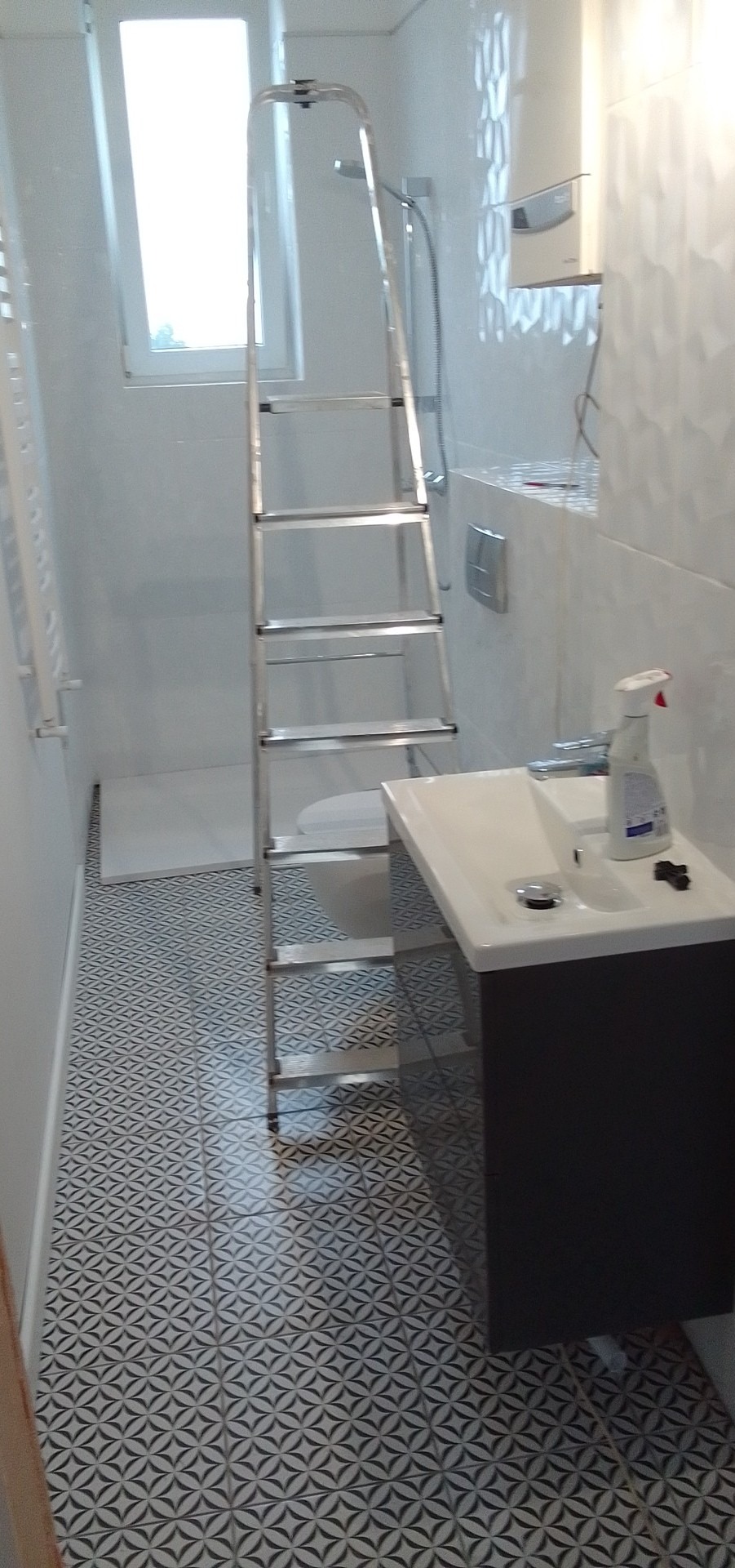 Łazienka w trakcie prac wykończeniowych: białe płytki 3D na ścianie, wzorzysta posadzka, drabina aluminiowa, częściowo widoczna toaleta i umywalka z szafką.