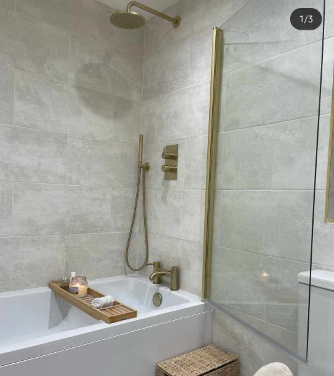 Nowoczesna łazienka z wanną, szklanym parawanem, złotą armaturą i szarymi płytkami, z drewnianą półką na wannie z świecą i ręcznikiem.