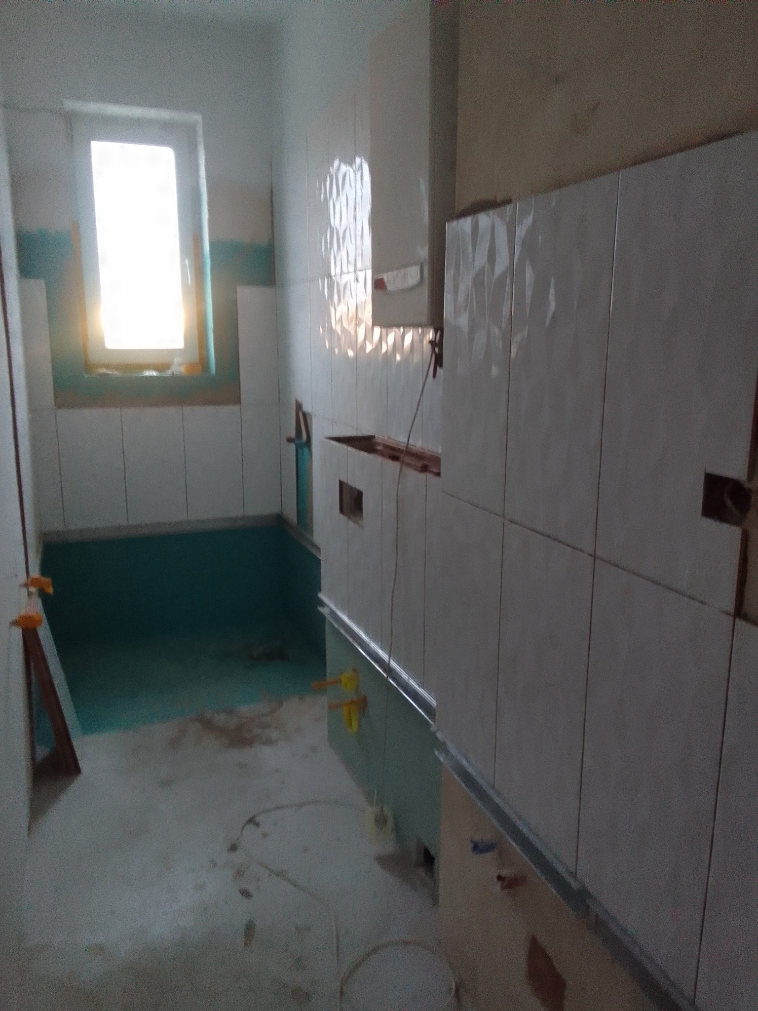 Łazienka w trakcie remontu, widoczne białe płytki z trójwymiarowym wzorem na ścianach, zabezpieczona folią podłoga, okno z widokiem na zewnątrz, elementy instalacji hydraulicznej wystające ze ściany.