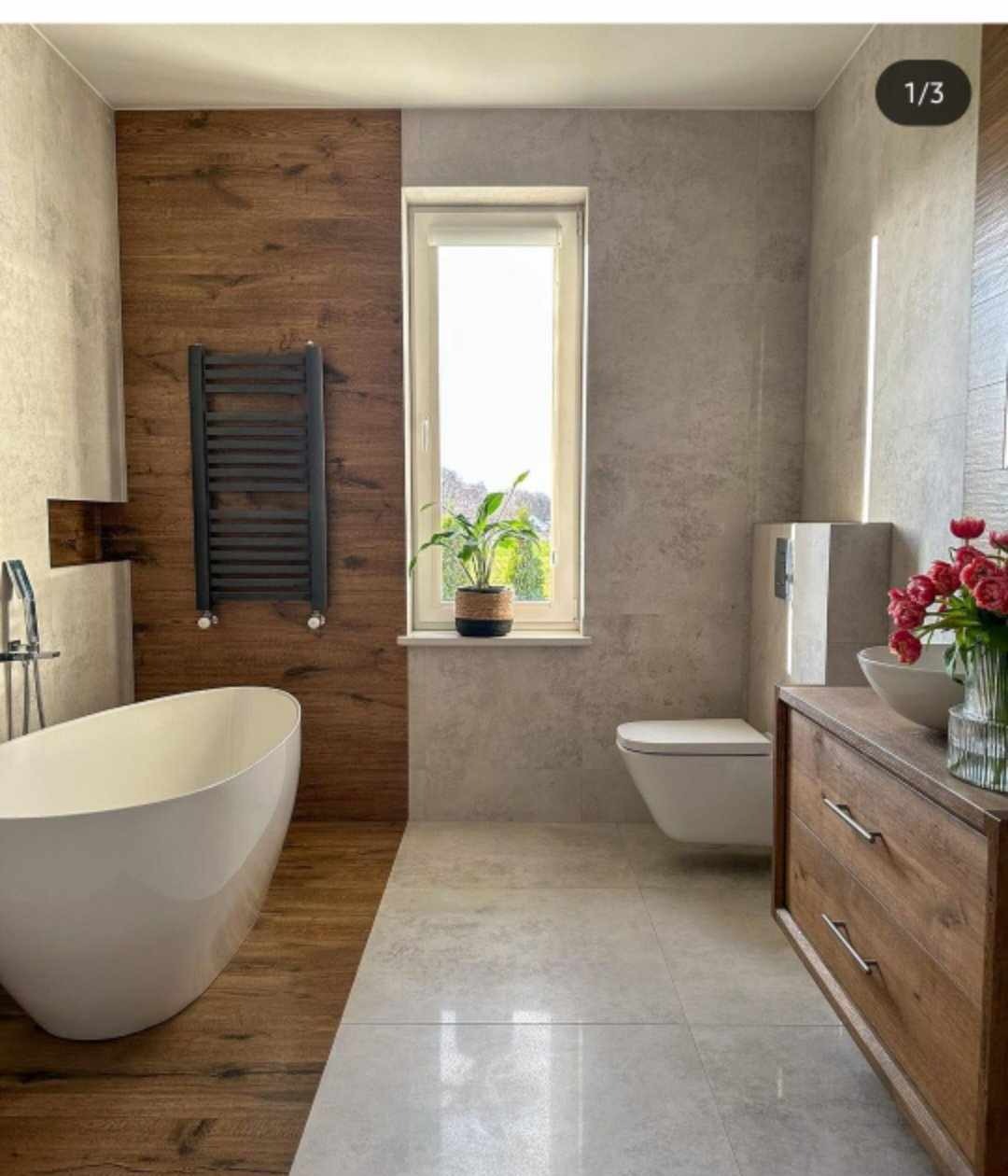 Nowoczesna łazienka z wolnostojącą wanną, drewnianą ścianą z grzejnikiem, szarymi płytkami, wiszącą toaletą i drewnianą szafką z kwiatami.