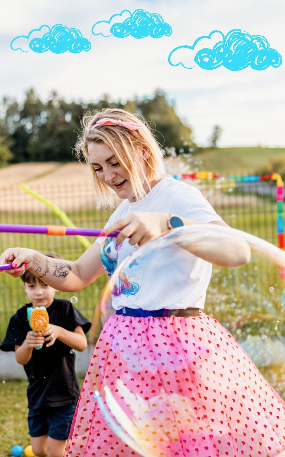 Kobieta w różowej tiulowej spódnicy i koszulce z jednorożcem puszcza duże bańki mydlane za pomocą specjalnych kijków, obok stoi chłopiec z pistoletem do baniek.