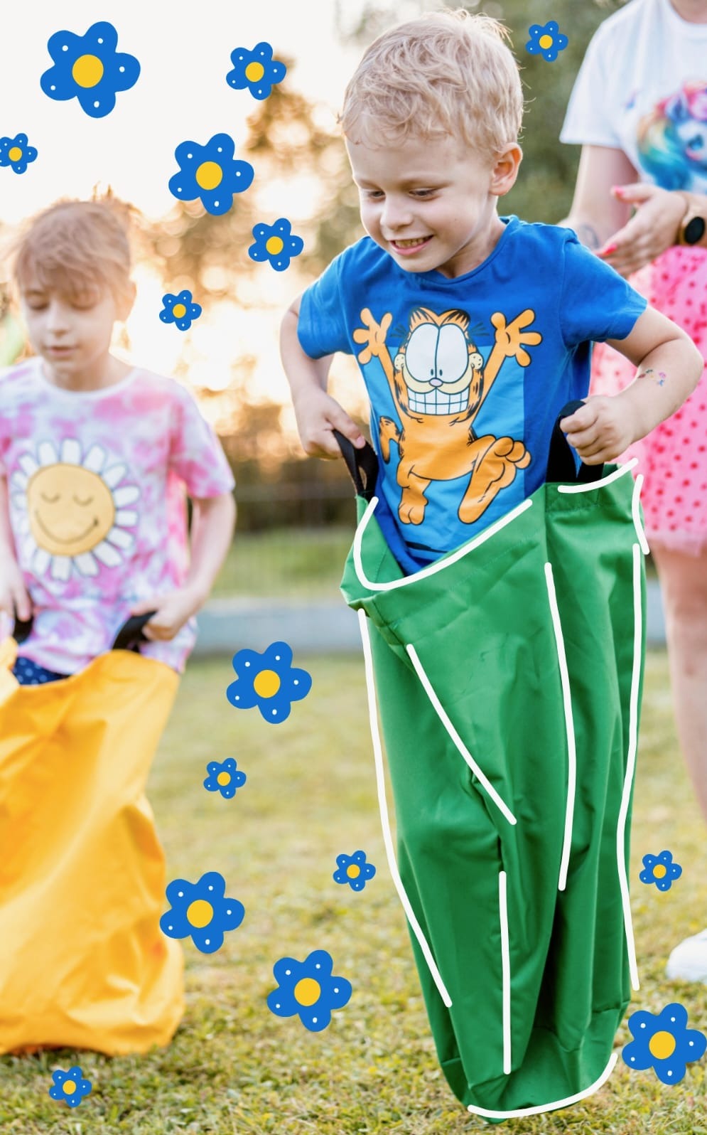 Dwójka dzieci w różnym wieku, biorąca udział w wyścigu w workach na trawie, z dekoracyjnymi niebieskimi kwiatkami w tle.