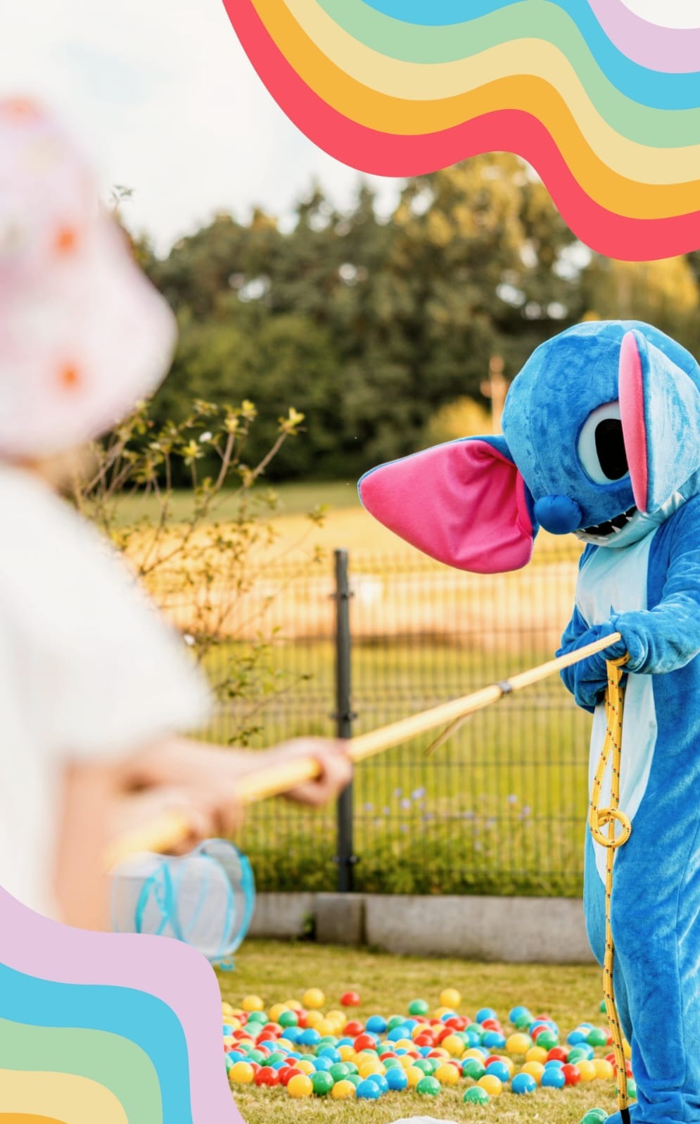 Animator w kostiumie Stitcha pomaga dziecku łowić kolorowe piłki z suchego basenu na trawie, w tle ogrodzenie i drzewa.