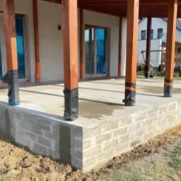 Wzniesiony fundament pod taras domu z drewnianymi słupami zabezpieczonymi folią, widoczne schody wejściowe i częściowo zagospodarowany teren.