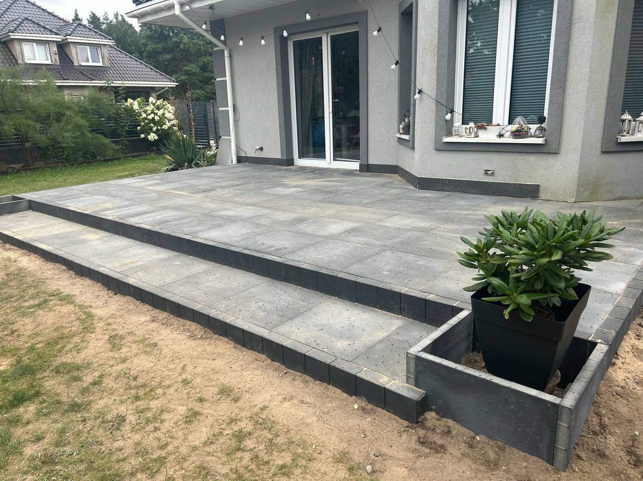 Wykończony taras z szarych płyt betonowych z dwoma stopniami, obrzeża z ciemnoszarego bruku, donica z rośliną, dom w tle, oświetlenie zewnętrzne.