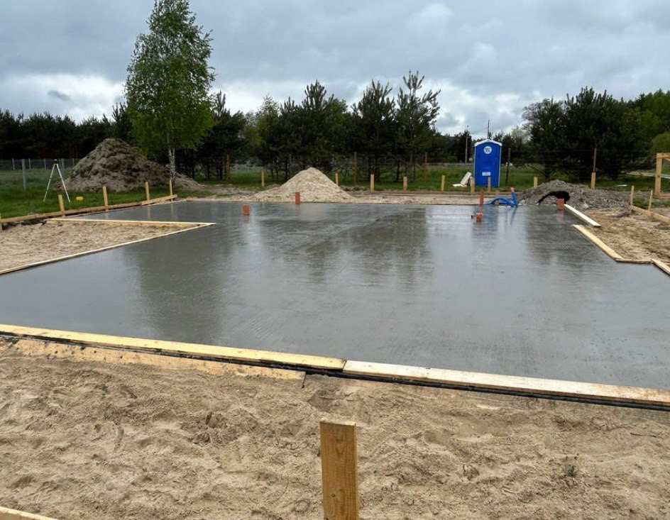 Zalana betonem płyta fundamentowa z drewnianym szalunkiem, widoczne wystające rury instalacyjne, w tle pryzma piasku i przenośna toaleta.